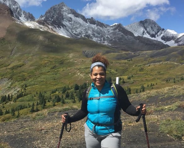 A black woman wearing a turquoise colored vest smiles at the camera. She is standing in a high mountain valley with a hiking pole in each hand and sharp, snow-covered mountain peaks behind her.