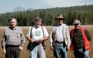 Four mature men dressed in jeans and T-shirts stand side by side in a large meadow with forest trees in the background.