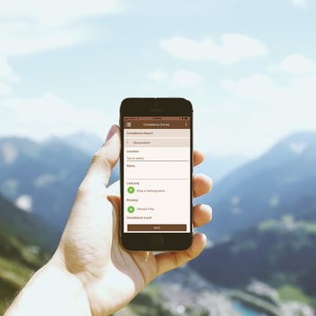 Hand holding a smartphone displaying an image of the Wildnote Mobile App on the screen. Mountains and a valley are in the background.